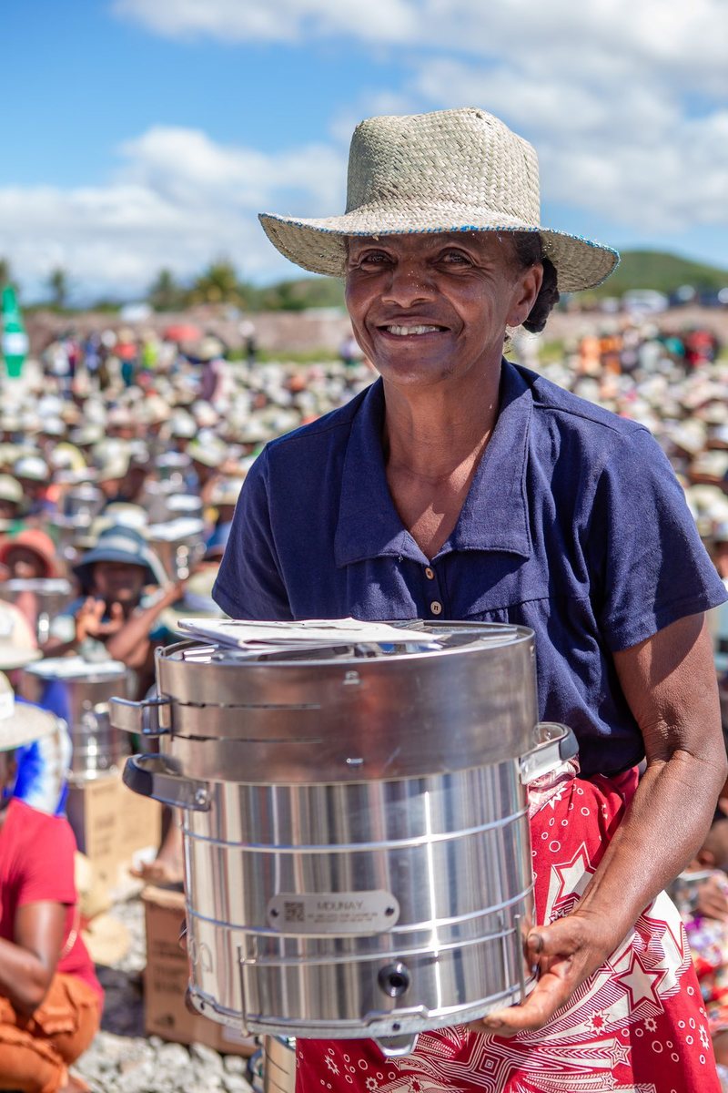 A smiling woman in a straw hat holds a stainless steel cookstove in front of a large seated crowd at a stove distribution event.